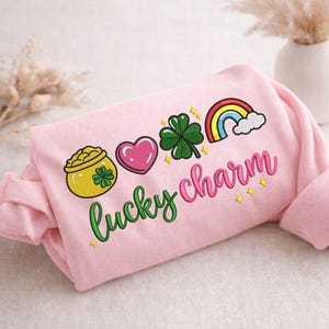 May include: A light pink sweatshirt with embroidered St. Patrick's Day themed graphics. The design includes a pot of gold, a pink heart, a green shamrock, and a rainbow. The words "lucky charm" are embroidered in a cursive font.