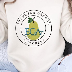 Könnte beinhalten: Cremefarbenes Sweatshirt mit einem gestickten kreisförmigen Design. Das Design enthält den Text "SOUTHERN OREGON" und "STITCHERS" um eine grüne Birne mit den Buchstaben "EGA" in Blau. Ein Blatt und ein Stiel befinden sich oben auf der Birne.