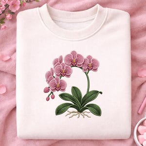 May include: A white sweatshirt featuring a detailed illustration of a pink orchid plant. The design includes the flower, leaves, and roots, with a vintage botanical style. The sweatshirt is folded on a pink surface.