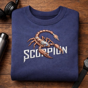 May include: Navy blue sweatshirt featuring a detailed scorpion design in shades of brown, tan, and silver, with the word "SCORPION" in white. The sweatshirt is folded on a wooden surface.