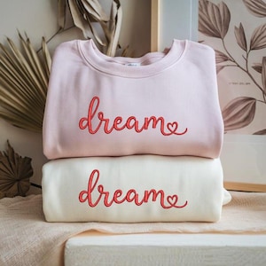 May include: Two sweatshirts, one pale pink and one cream, stacked on top of each other. Each sweatshirt has the word "dream" in red script with a heart. The sweatshirts are folded and appear soft.