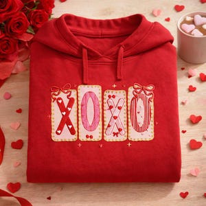 Könnte beinhalten: Ein gefalteter roter Hoodie mit den gestickten Buchstaben "XOXO" in einem dekorativen Stil. Die Buchstaben sind rosa und weiß, mit goldenen Verzierungen und Schleifen. Der Hoodie ist von Valentinstagsdekorationen umgeben.