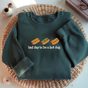 May include: A dark green sweatshirt with three embroidered hot dogs and the phrase "bad day to be a hot dog" in white. The sweatshirt is folded on a woven mat.