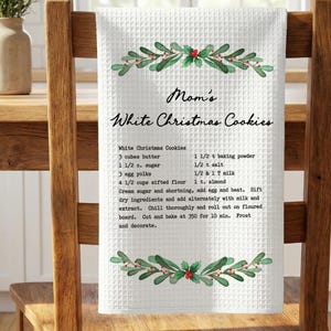 May include: White waffle-weave kitchen towel with a recipe for "Mom's White Christmas Cookies." The towel has a decorative border of green leaves and red berries. The recipe includes butter, sugar, eggs, and flour.