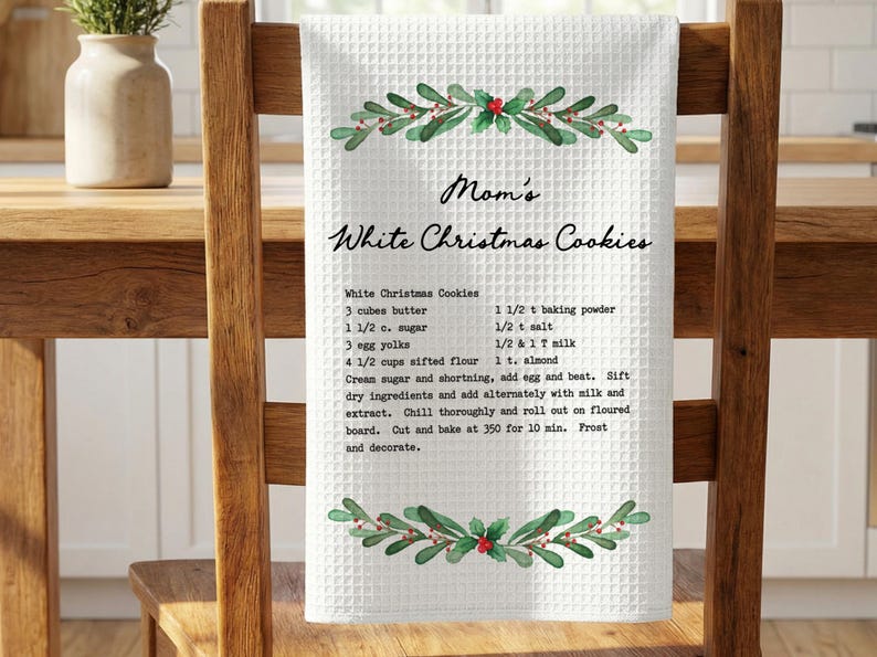 May include: White waffle-weave kitchen towel with a recipe for "Mom's White Christmas Cookies." The towel has a decorative border of green leaves and red berries. The recipe includes butter, sugar, eggs, and flour.