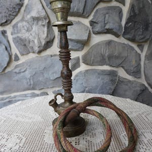 May include: An antique brass hookah with a decorative, multi-colored hose. The hookah has a tall, ornate stem and a small spout. The hose is wrapped in a patterned fabric with red, green, and brown hues. The items are on a white lace tablecloth.