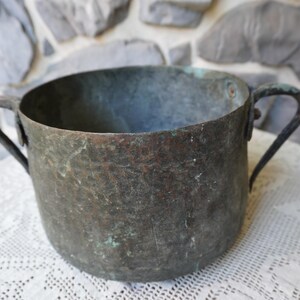 May include: A weathered, antique copper pot with two curved handles. The pot has a dark, mottled patina with hints of green and brown, suggesting age and use. It sits on a white lace tablecloth against a stone background.
