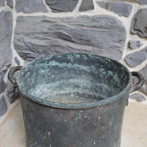 May include: A large, aged copper cauldron with two side handles. The metal has a dark, weathered appearance with areas of green oxidation, suggesting outdoor use. The cauldron sits on a light-colored surface against a stone wall.