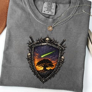 May include: Pepper-colored t-shirt with a shield graphic featuring a tree, a shooting star, and skulls. The shirt has a crew neck and short sleeves. A silver necklace with a pendant is draped across the shirt.