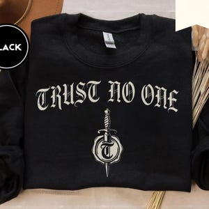 May include: Black sweatshirt featuring the phrase "TRUST NO ONE" in a decorative font, with a dagger and seal graphic below. The long-sleeved, crew-neck top has the word "BLACK" in a black circle in the upper left corner.