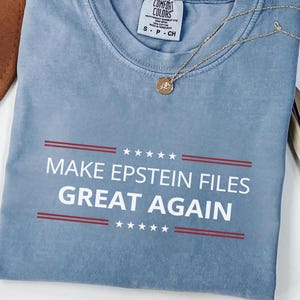 Może przedstawiać: Niebieska koszulka Comfort Colors z białym napisem "MAKE EPSTEIN FILES GREAT AGAIN", z czerwonymi liniami i gwiazdkami. Widoczny jest również złoty naszyjnik i brązowy kapelusz.