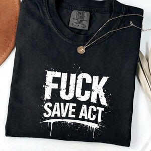 Peut inclure: T-shirt noir avec l'inscription blanche "FUCK SAVE ACT" en lettres délavées. Le t-shirt est plié et porte une petite étiquette sur le col. Un collier doré avec un pendentif est posé sur le t-shirt.