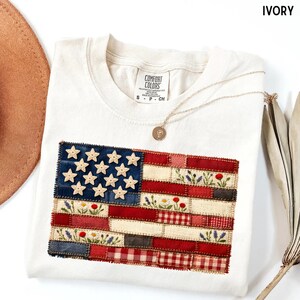 以下が含まれることがあります： アイボリー色のTシャツ。刺繍されたアメリカ国旗のデザインが特徴です。星型のアクセントが付いた青いフィールド、赤いストライプと白いストライプ、花の刺繍が施されています。ペンダント付きのゴールドネックレスがTシャツの上にかけられています。