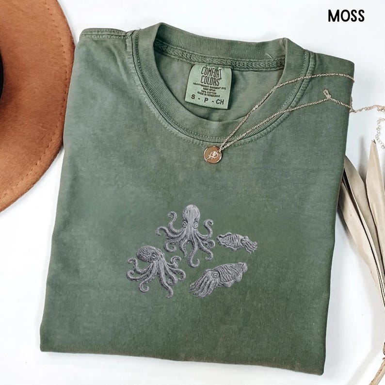 May include: Moss green t-shirt with embroidered gray octopus and squid designs. The shirt has a crew neck and short sleeves. A gold necklace with a pendant is draped across the shirt. The tag reads "Comfort Colors".