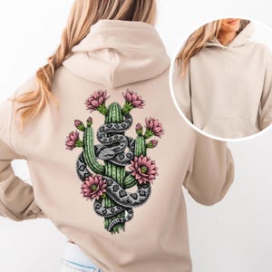 Può includere: Felpa con cappuccio beige con grafica sul retro raffigurante un cactus verde, fiori rosa e un serpente grigio e nero avvolto attorno al cactus. La felpa con cappuccio ha una tasca frontale e un cappuccio. Lo sfondo è beige chiaro.