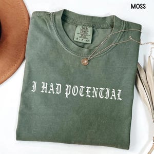 Puede incluir: Una camiseta verde musgo con el texto "I HAD POTENTIAL" en fuente gótica blanca. Un collar dorado con un colgante descansa sobre la camiseta. La camiseta es de la marca Comfort Colors, talla S-P-CH.