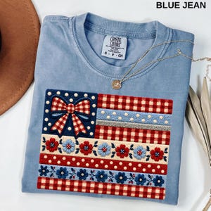 May include: A light blue t-shirt with an embroidered American flag design. The flag features a red and white gingham pattern, blue polka dots, and floral accents. The shirt has the text "BLUE JEAN" at the top.