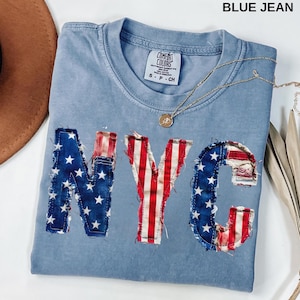 Könnte beinhalten: Hellblaues T-Shirt mit den Buchstaben "NYC" auf der Vorderseite in einem patriotischen Design. Das "N" und "C" sind blau mit weißen Sternen, und das "Y" ist rot und weiß gestreift. Eine goldene Halskette mit Anhänger ist sichtbar.