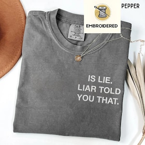 May include: A grey Comfort Colors t-shirt with the embroidered text "IS LIE. LIAR TOLD YOU THAT." An embroidered logo is in the upper right corner. A silver necklace with a pendant is draped across the shirt.