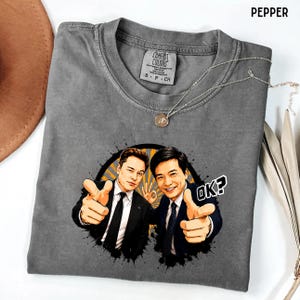 Puede incluir: Camiseta gris Comfort Colors con un gráfico de dos hombres señalando al espectador y el texto "OK?". Un collar plateado con un colgante está sobre la camiseta. Talla S-P-CH.
