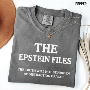 Könnte beinhalten: Ein graues T-Shirt mit dem weißen Text "THE EPSTEIN FILES" und dem Satz "THE TRUTH WILL NOT BE HIDDEN BY DISTRACTION OR WAR." Eine silberne Halskette mit Anhänger liegt auf dem Shirt. Das Shirt ist mit "PEPPER" gekennzeichnet.