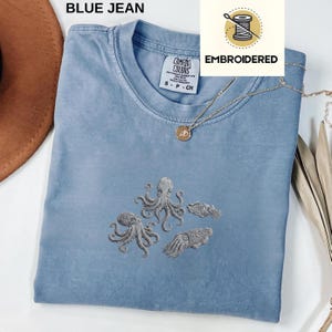 May include: A blue jean Comfort Colors t-shirt with embroidered gray octopus and squid designs. The shirt has a crew neck and short sleeves. A gold necklace with a pendant is draped on the shirt. The word "EMBROIDERED" is printed on a white background.