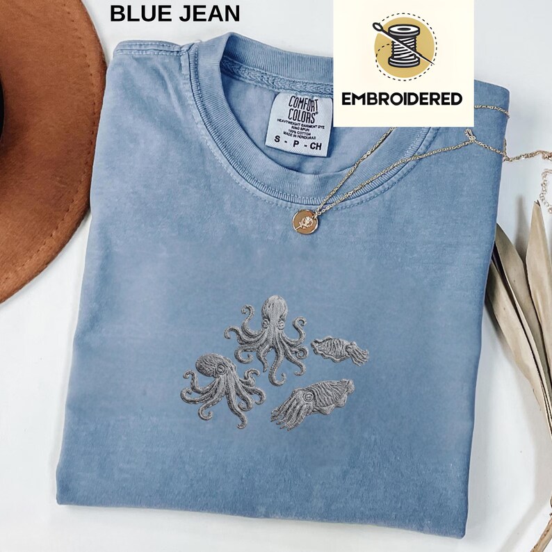 May include: A blue jean Comfort Colors t-shirt with embroidered gray octopus and squid designs. The shirt has a crew neck and short sleeves. A gold necklace with a pendant is draped on the shirt. The word "EMBROIDERED" is printed on a white background.