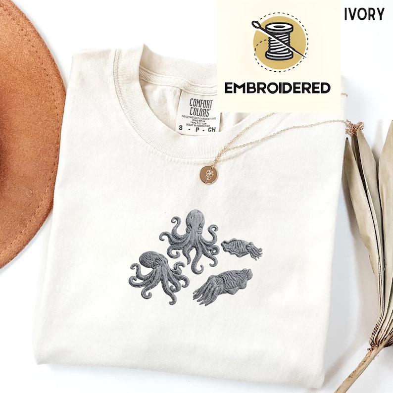 May include: Ivory-colored t-shirt with embroidered gray octopus and cuttlefish designs. The shirt has a crew neck and short sleeves. The text "EMBROIDERED" is above the design. A gold necklace with a pendant is also visible.