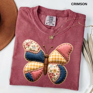Könnte beinhalten: Karmesinrotes T-Shirt mit einem gestickten Schmetterlingsdesign. Der Schmetterling hat Patchwork-Flügel in floralen, Jeans-, rosa Cord- und gelb-karierten Mustern. Eine goldene Halskette mit Anhänger liegt auf dem Shirt.