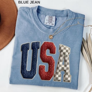 Pode incluir: Camiseta azul clara com a palavra "USA" em letras azul marinho, vermelhas e xadrez cinza. A camiseta tem gola redonda e uma pequena etiqueta. Um colar de ouro com um pingente está drapeado sobre a camiseta. O texto "BLUE JEAN" está no topo.