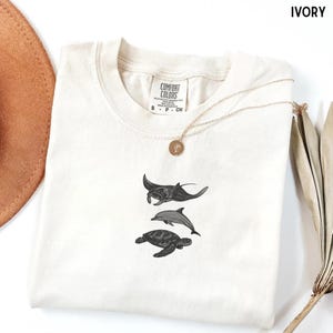 以下が含まれることがあります： マンタ、イルカ、ウミガメのデザインが施されたアイボリー色のTシャツ。 シャツはクルーネックと半袖です。 ペンダント付きのゴールドネックレスがシャツの上にかけられています。 シャツにはComfort Colorsのタグが付いています。