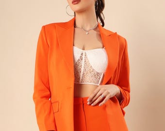 Könnte beinhalten: Helles orangefarbenes Blazer- und Hosen-Ensemble. Der Blazer hat ein klassisches Revers und einen Zwei-Knopf-Verschluss. Die hoch taillierte Hose hat ein gerades Bein. Das Outfit wird durch ein weißes Spitzentop ergänzt. Ein modisches und modernes Outfit.