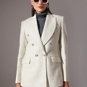 May include: Cream-colored double-breasted blazer with silver buttons. The blazer has a textured weave and is paired with matching trousers. The outfit is worn with a gray turtleneck and sunglasses.