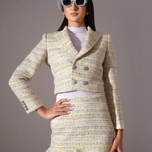 May include: A yellow, white, and gray tweed skirt suit. The double-breasted jacket has silver buttons and the skirt has a ruffled hem. The person is wearing a white top, sunglasses, and a gray beret.