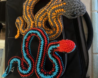 Peut inclure: Deux serpents crochetés. L'un est gris et orange, l'autre rouge, noir et bleu. Les serpents sont posés sur une surface noire. Les serpents sont faits à la main et ont un aspect doux et texturé. Ils sont probablement des jouets ou des objets décoratifs.