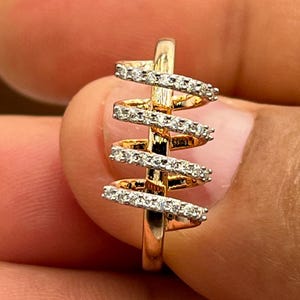 May include: A gold-toned ring with a spiral design. The ring features multiple horizontal bands encrusted with small, sparkling gemstones. The ring is held between fingers, showcasing its intricate details and craftsmanship.