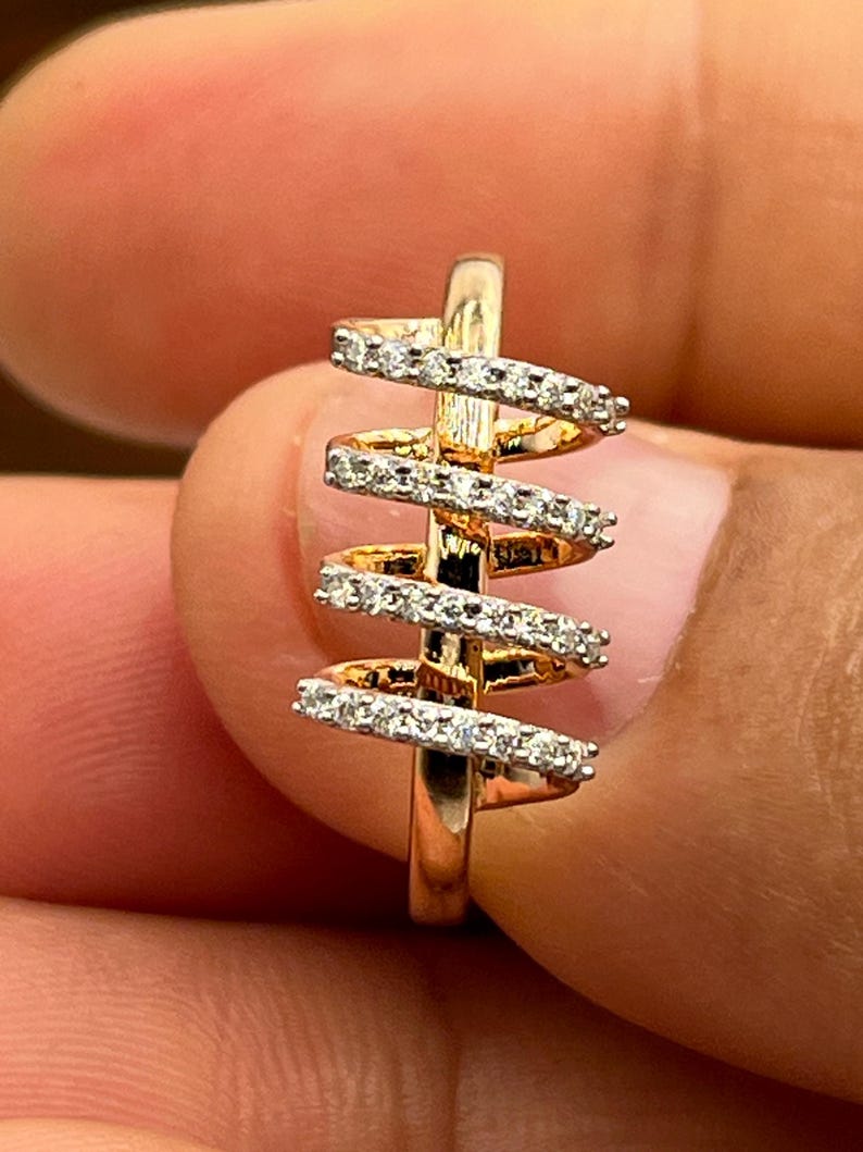 May include: A gold-toned ring with a spiral design. The ring features multiple horizontal bands encrusted with small, sparkling gemstones. The ring is held between fingers, showcasing its intricate details and craftsmanship.