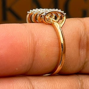 May include: A gold ring with a cluster of small, clear gemstones. The ring has a delicate band and a multi-layered design around the gemstones. The ring is shown on a finger, highlighting its intricate details and sparkle.