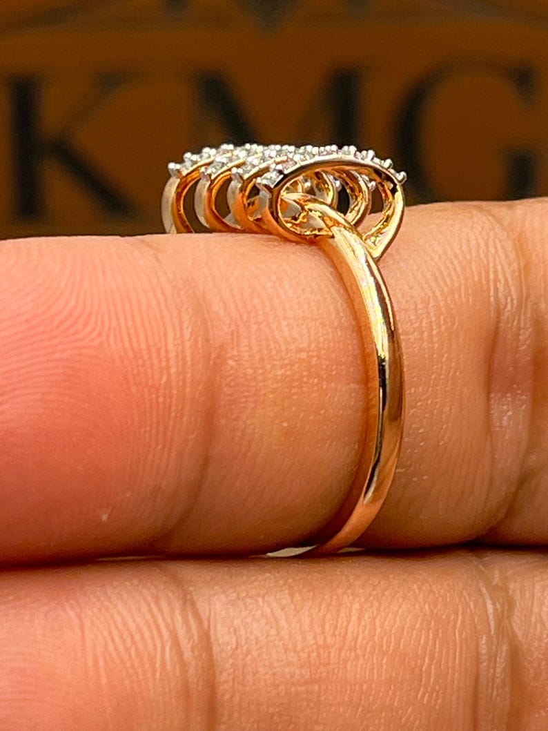 May include: A gold ring with a cluster of small, clear gemstones. The ring has a delicate band and a multi-layered design around the gemstones. The ring is shown on a finger, highlighting its intricate details and sparkle.