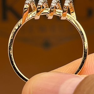 May include: A gold-colored ring with a unique design. The ring features four vertical, curved elements, each adorned with small, sparkling stones. The ring is held by a finger, showcasing its intricate details and craftsmanship. The ring's band is a simple, polished gold.