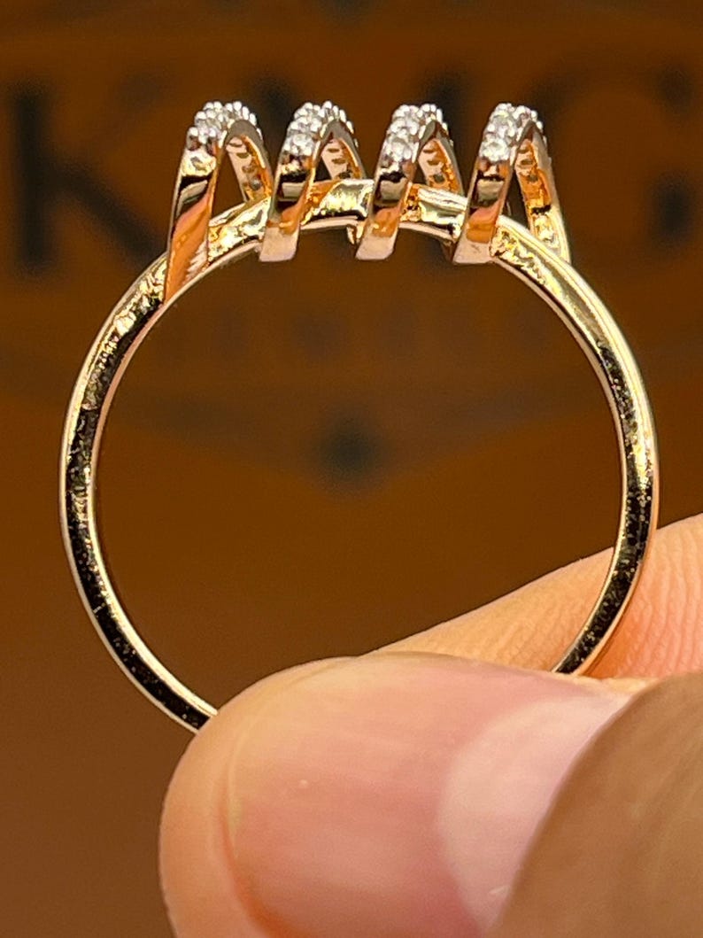 May include: A gold-colored ring with a unique design. The ring features four vertical, curved elements, each adorned with small, sparkling stones. The ring is held by a finger, showcasing its intricate details and craftsmanship. The ring's band is a simple, polished gold.