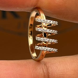 May include: A gold-toned ring with a unique design. The ring features a series of horizontal bars adorned with small, sparkling stones, creating a spiral effect. The band is smooth and polished, and the ring is held by a hand.