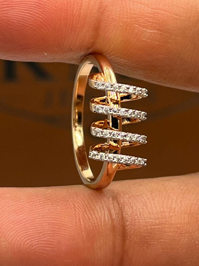 May include: A gold-toned ring with a unique design. The ring features a series of horizontal bars adorned with small, sparkling stones, creating a spiral effect. The band is smooth and polished, and the ring is held by a hand.