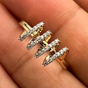 May include: A gold-toned ring with a unique design. The ring features three X-shaped elements, each encrusted with small, sparkling white gemstones. The ring is held in a hand, showcasing its intricate details and craftsmanship.