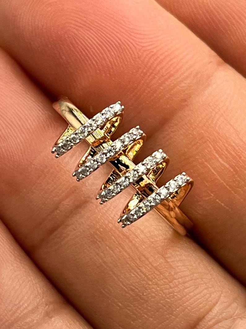 May include: A gold-toned ring with a unique design. The ring features three X-shaped elements, each encrusted with small, sparkling white gemstones. The ring is held in a hand, showcasing its intricate details and craftsmanship.