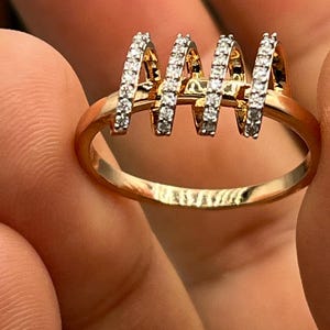 May include: A gold-toned ring with four vertical, arched sections encrusted with small, clear gemstones. The ring's band is smooth and rounded, and the gemstones sparkle against the gold.