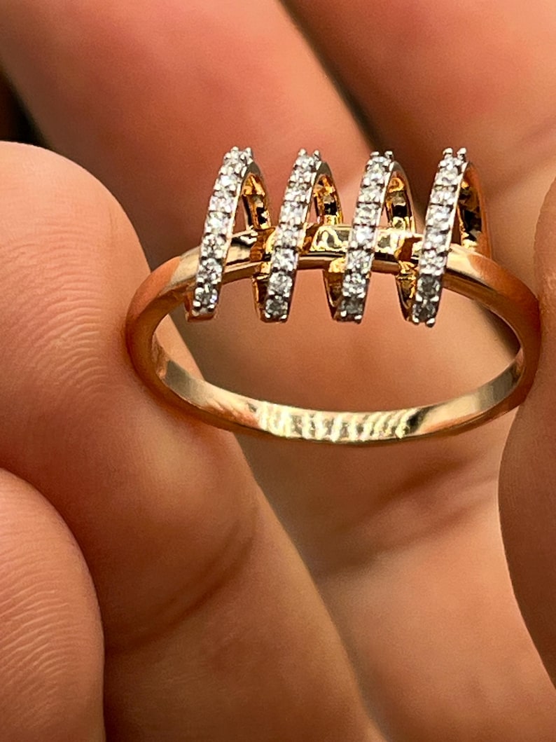 May include: A gold-toned ring with four vertical, arched sections encrusted with small, clear gemstones. The ring's band is smooth and rounded, and the gemstones sparkle against the gold.