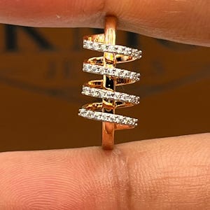 May include: A rose gold ring with a unique spiral design. The ring features multiple bands of small, sparkling white stones set along the spiral. The ring is held between two fingers.