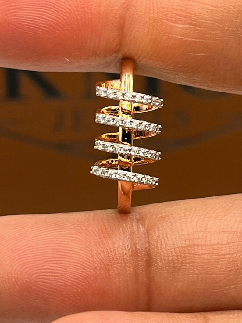May include: A rose gold ring with a unique spiral design. The ring features multiple bands of small, sparkling white stones set along the spiral. The ring is held between two fingers.