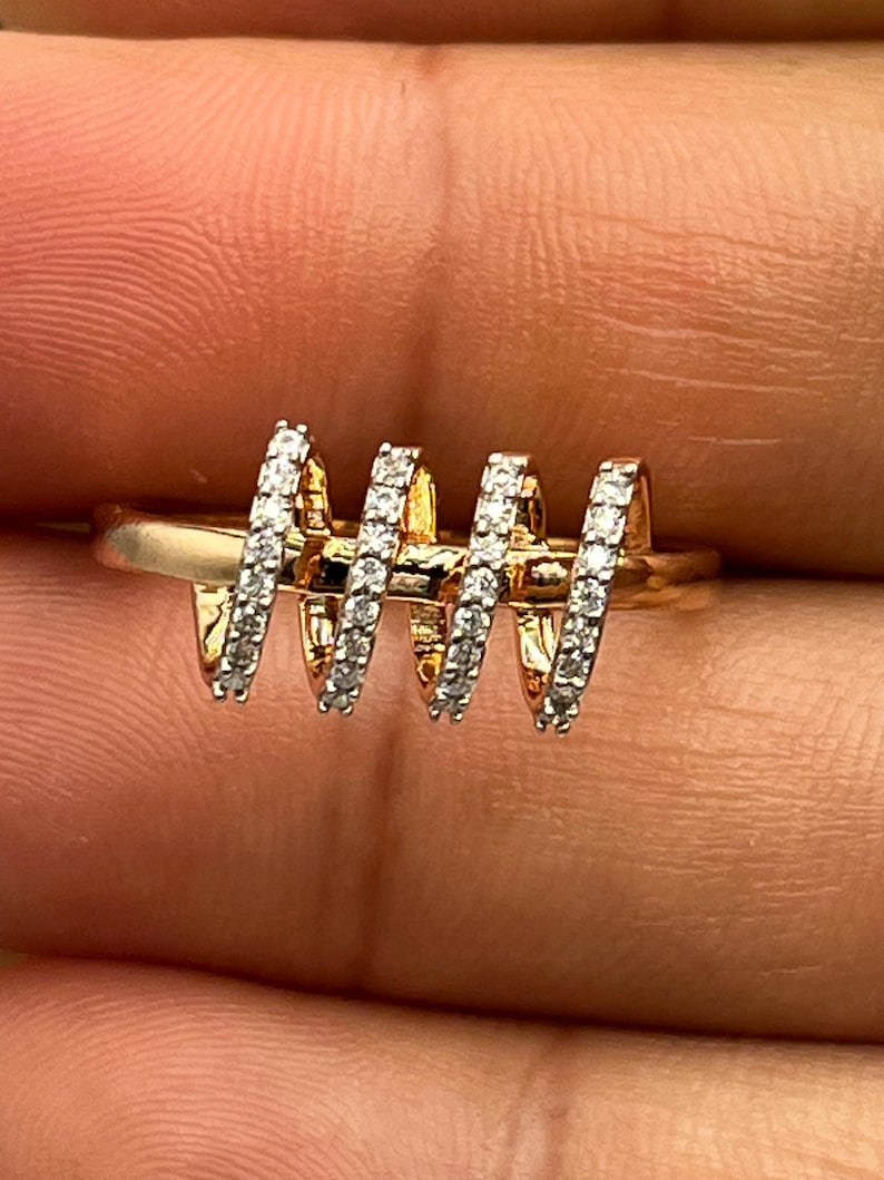 May include: A gold-toned ring with a unique design. The ring features four spiraling sections, each encrusted with small, sparkling stones. The ring is set against a neutral background, highlighting its intricate details and craftsmanship.